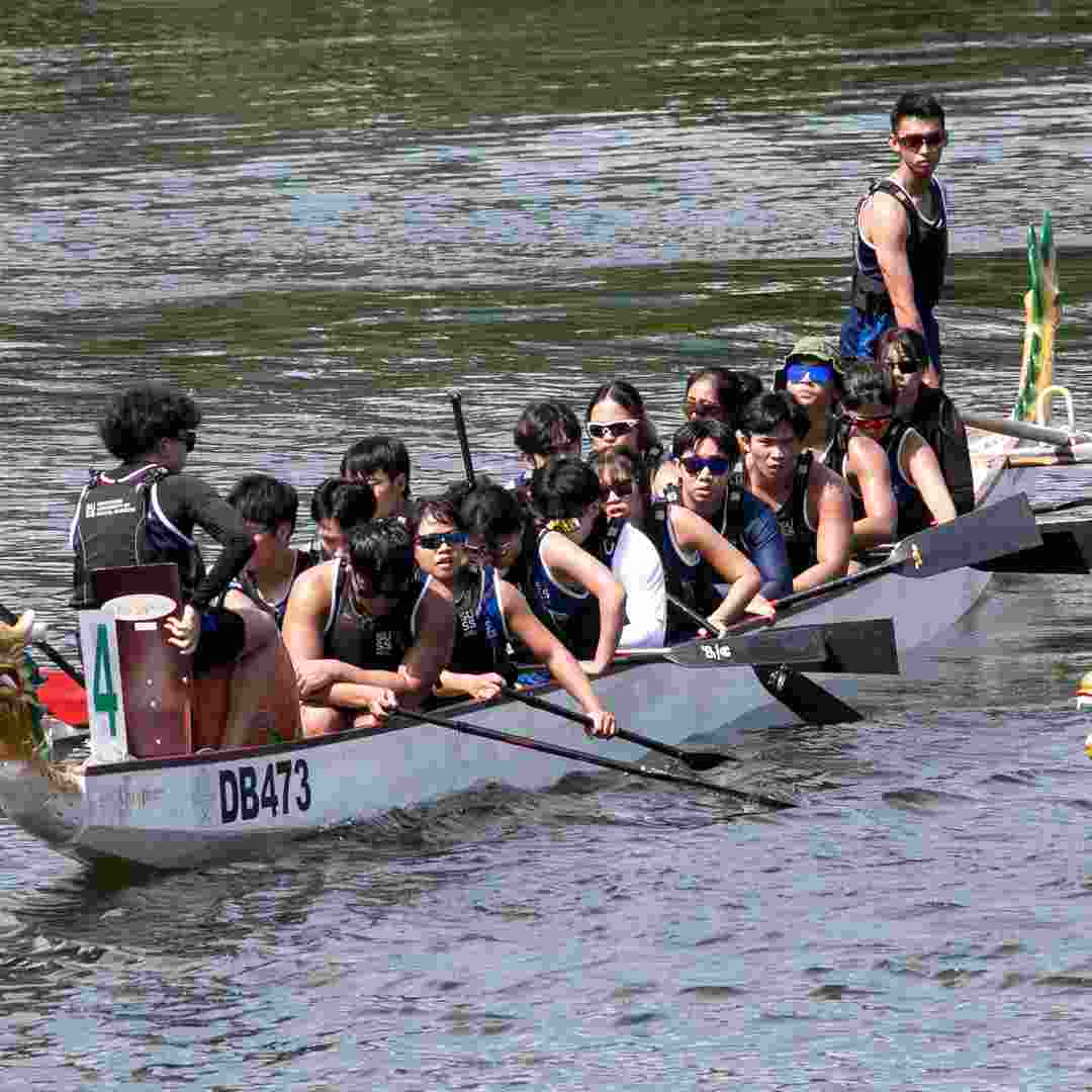 20 dragonboaters paddling in sync in the river with, a leader standing at the front of the boat, and the navigator standing at the end of the boat. 