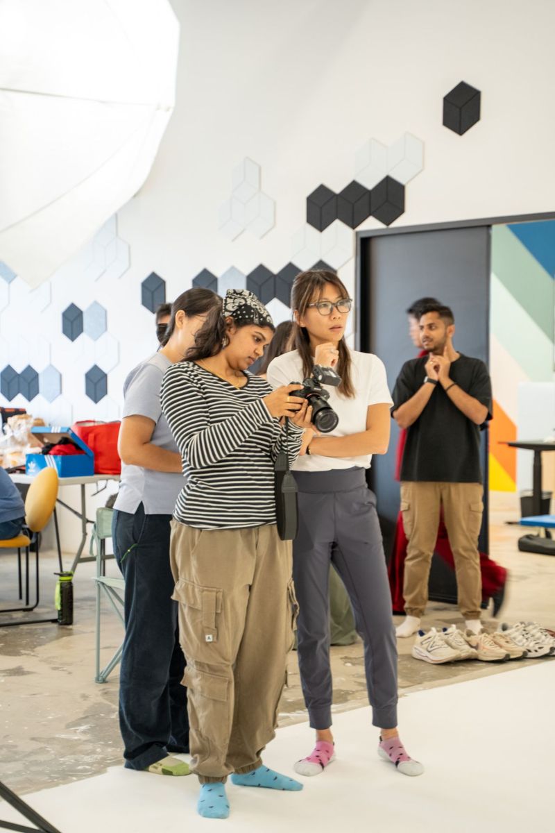 A group of individuals standing together in a room, smiling and wearing casual clothing such as jeans and footwear. The background features a wall and some furniture.