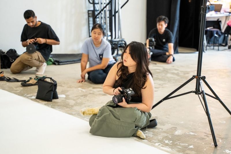 A group of photographers sits on the floor in a studio, focusing on a photoshoot while holding their cameras.