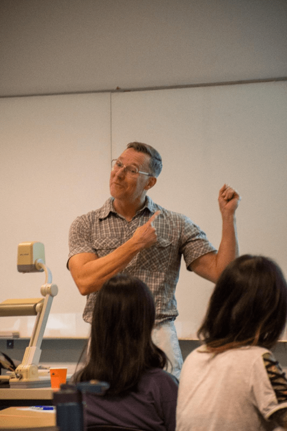 A man stands at the front of a classroom gesturing animatedly while speaking to students seated and facing him