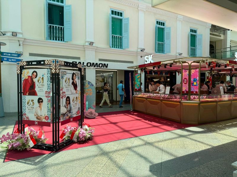 A vibrant shopping mall scene featuring a promotional display and a decorated stall, with people walking on a red carpet.