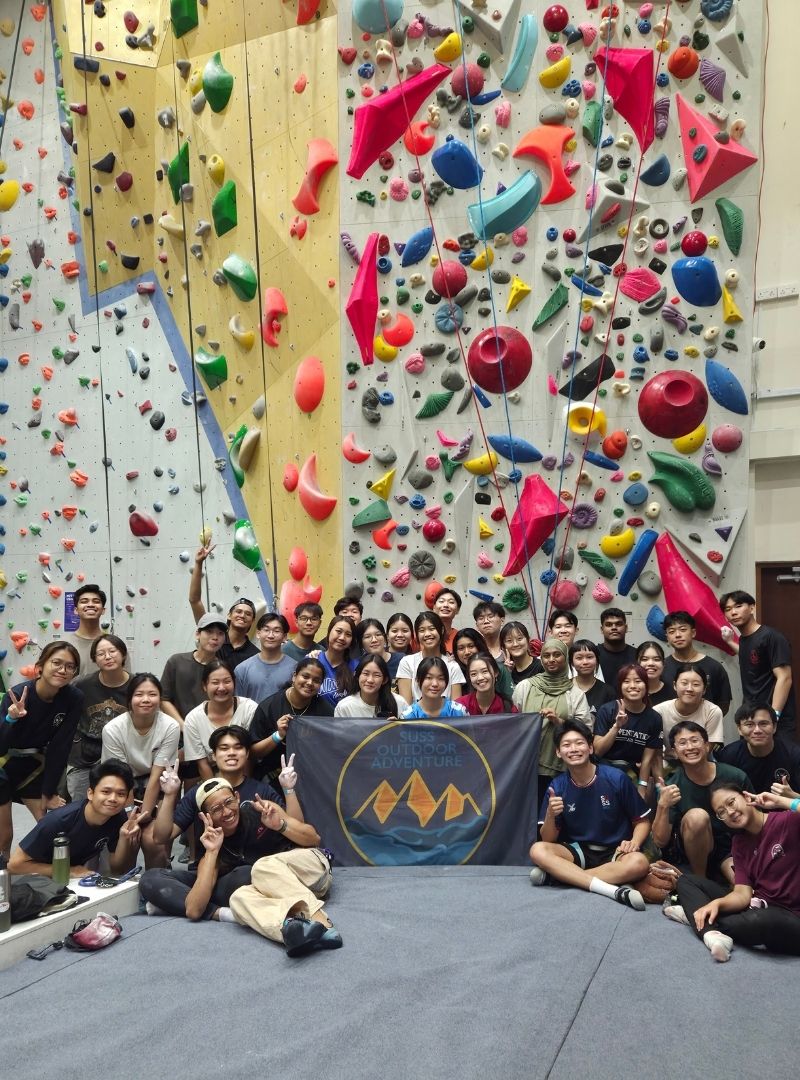 Participants, EXCO facilitators, and volunteer rock climbing members gather for a final group photo in front of the high wall at Outpost Climbing Gym, proudly holding the ODAIG flag.