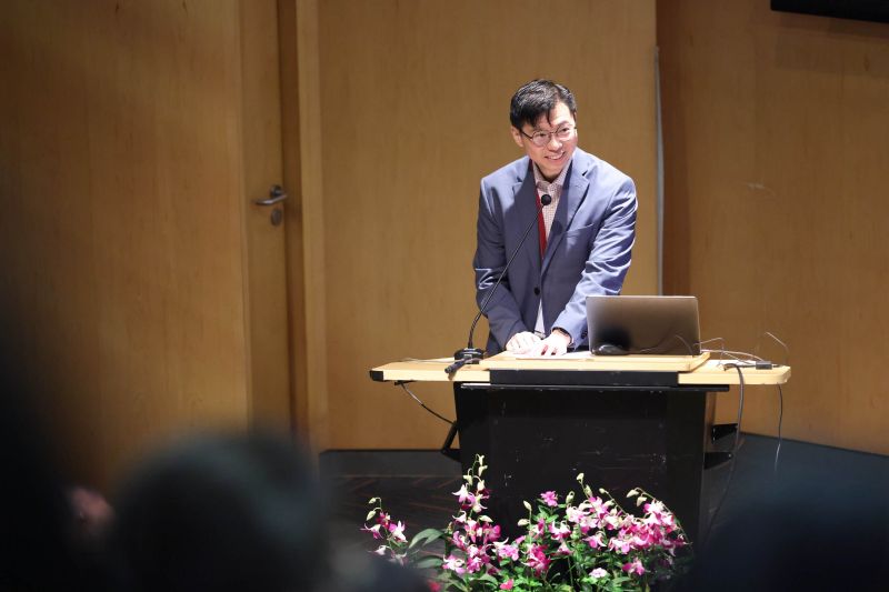 Person in a suit speaking at a podium with a laptop and flowers