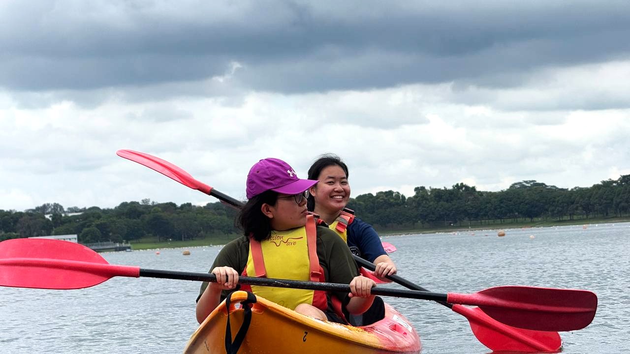From a front view of the kayak, Lim Kym Suan, Esther smiles warmly at the camera while the client takes a short break, both sharing a calm moment on the water.