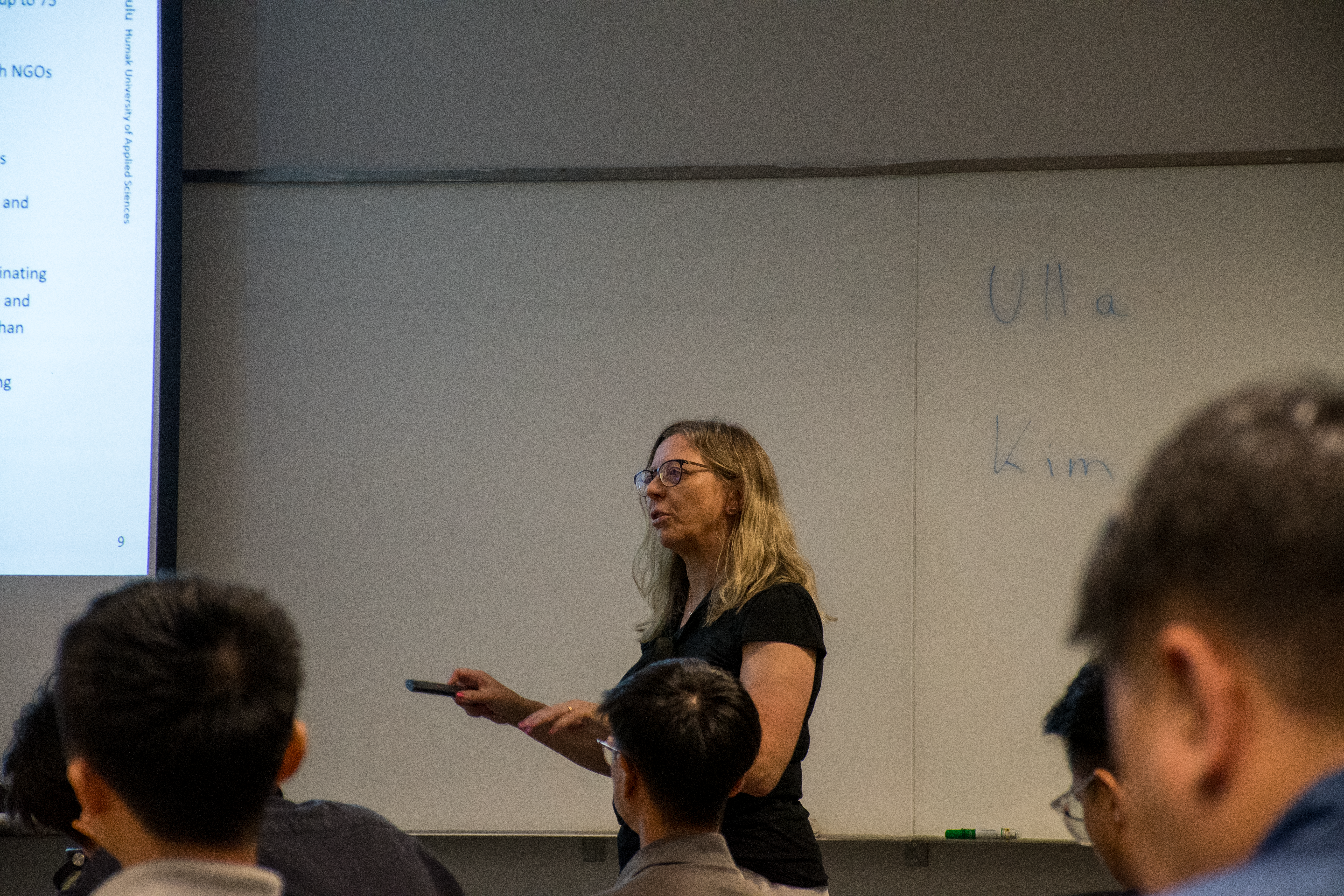 A presenter stands in front of a whiteboard, gesturing towards a projector screen while students attentively observe.