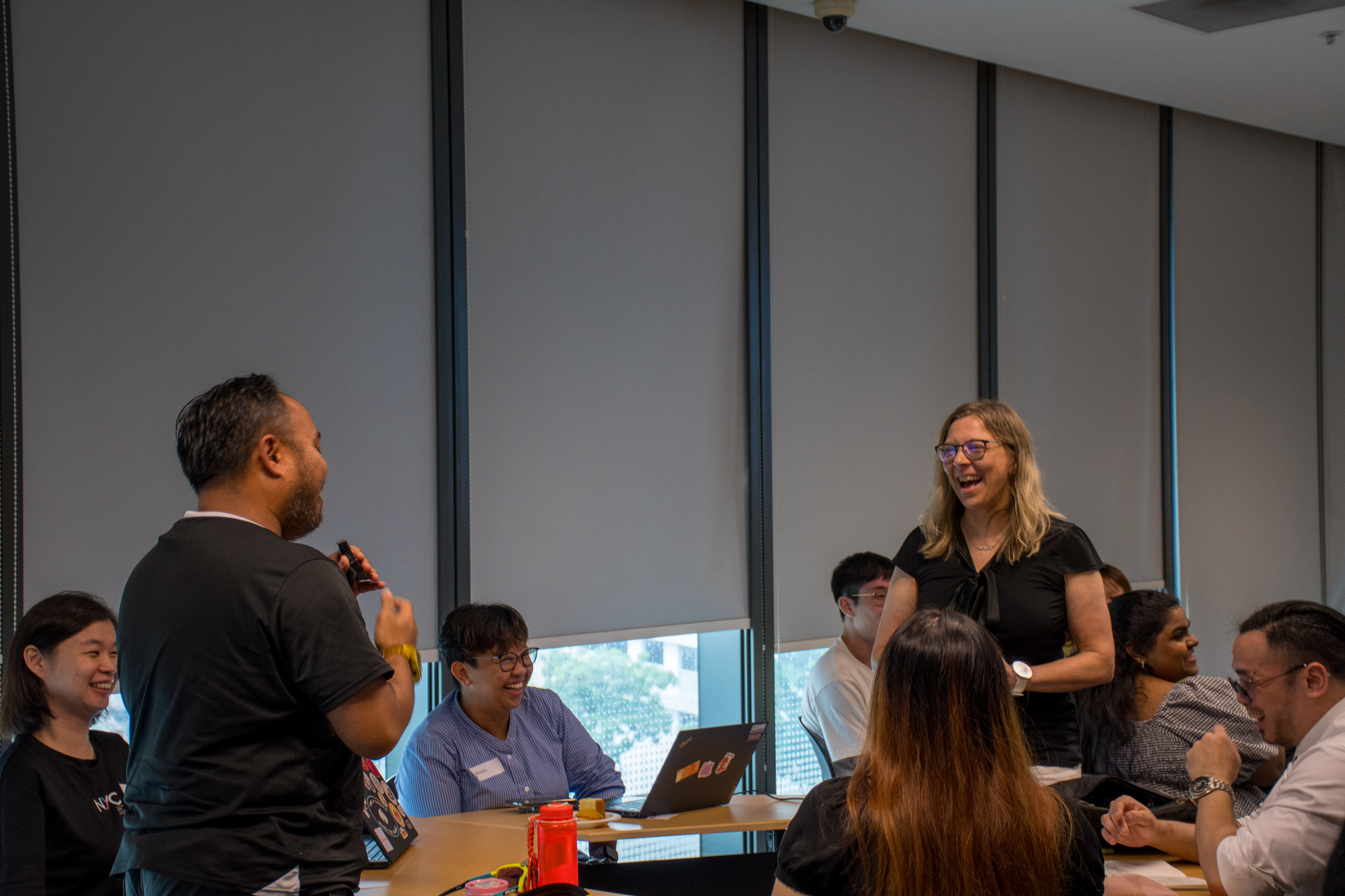 A group of people sit around a conference table while a man stands and speaks, and a woman standing nearby laughs. Others watch and smile in a modern office s