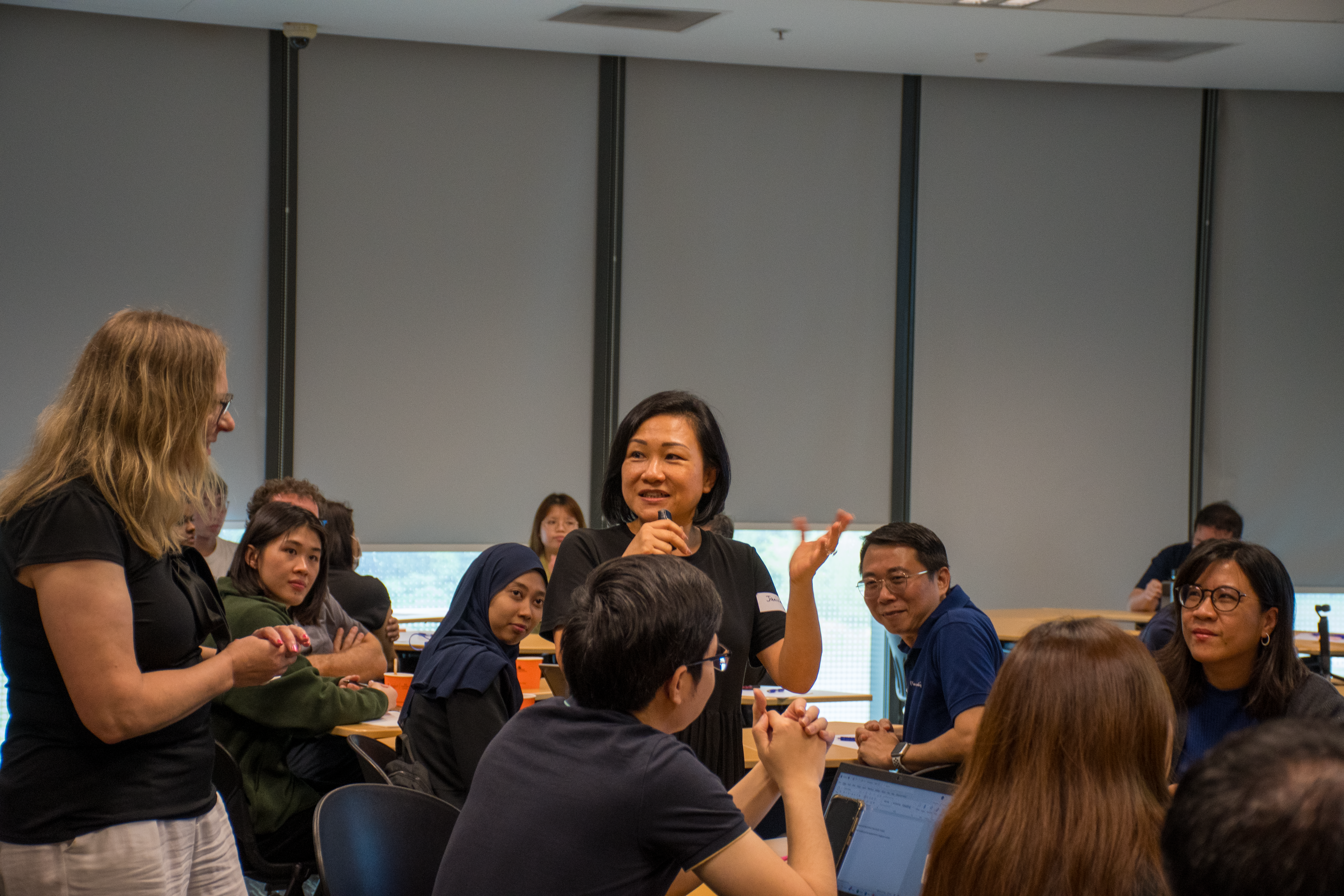 A group of people are engaged in conversation during a collaborative workshop in a well-lit classroom with large windows.
