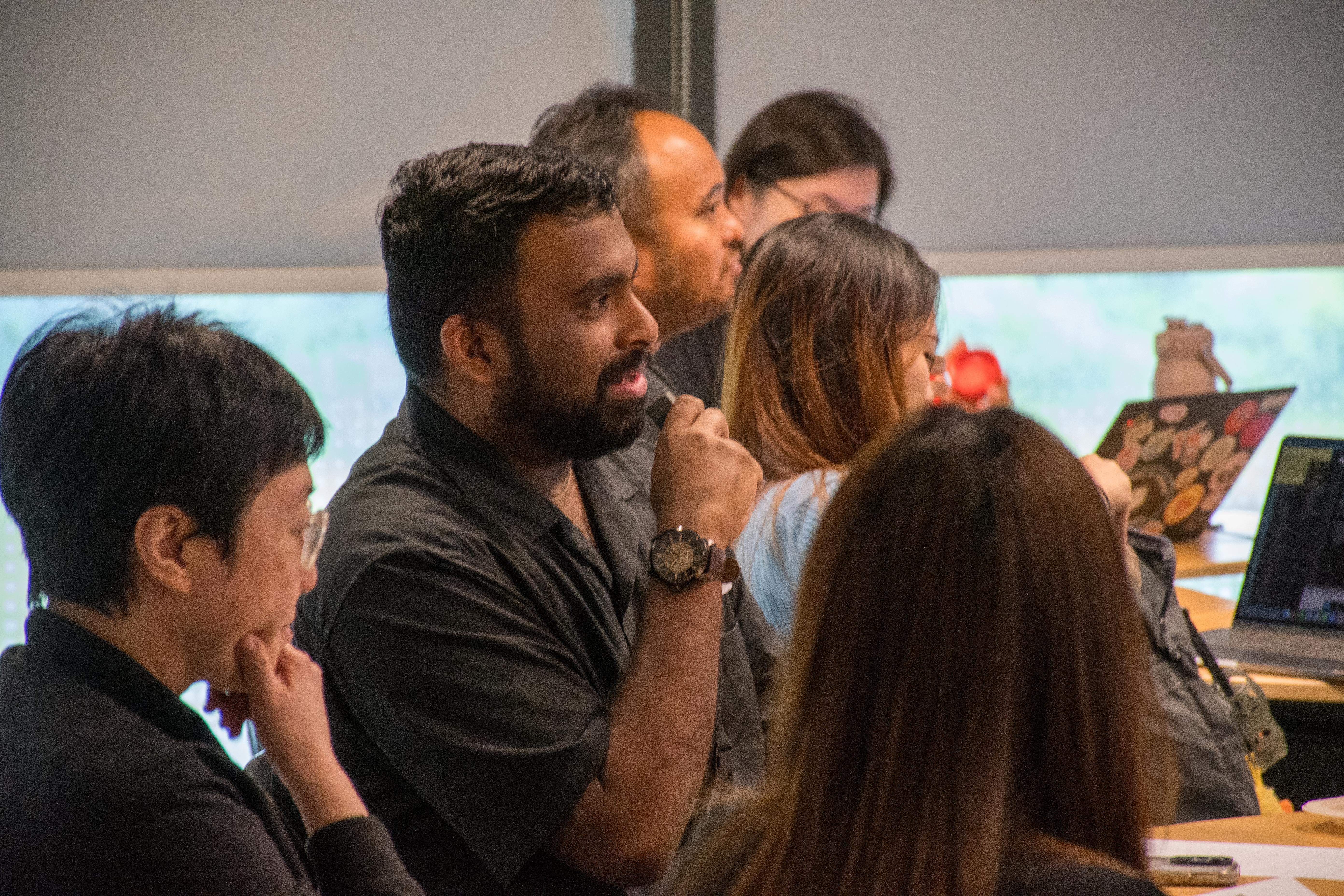 A group of people attentively listening in a modern classroom setting, with laptops and notebooks on their desks.