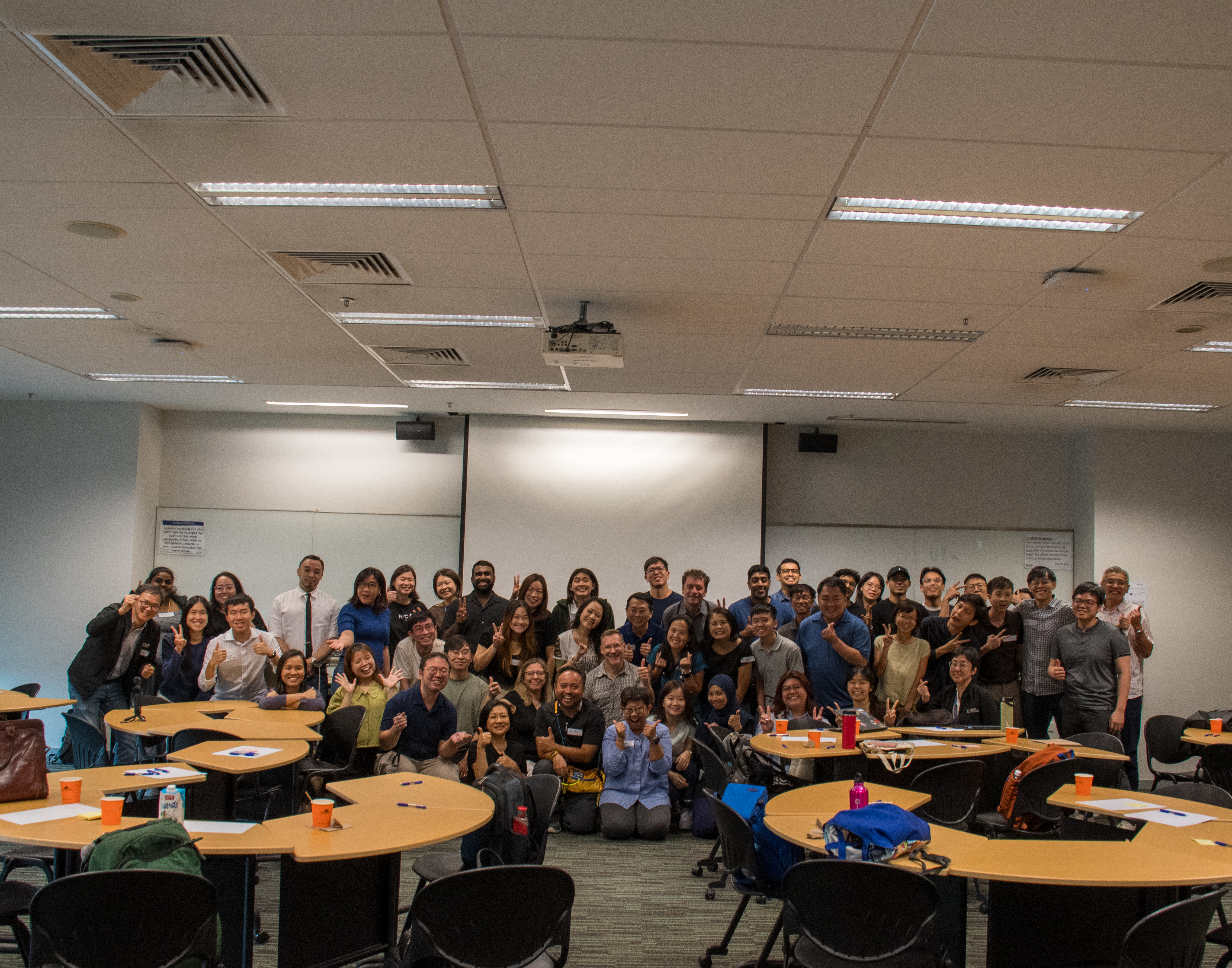 A large group of diverse people posing together in a classroom setting, smiling and making hand gestures, seated and standing around tables.