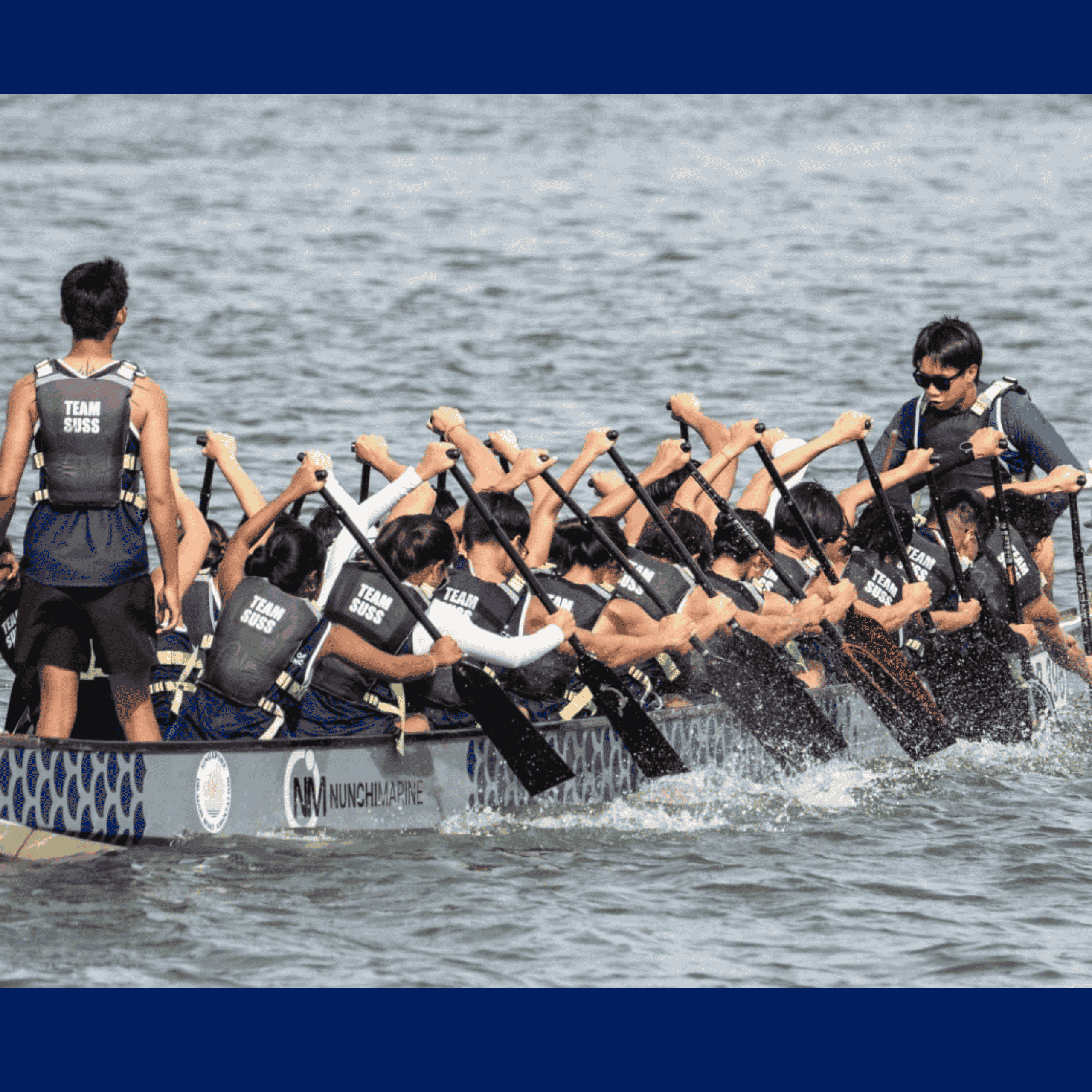 20 dragonboaters paddling in sync in the river with, a leader standing at the front of the boat, and the navigator standing at the end of the boat. 